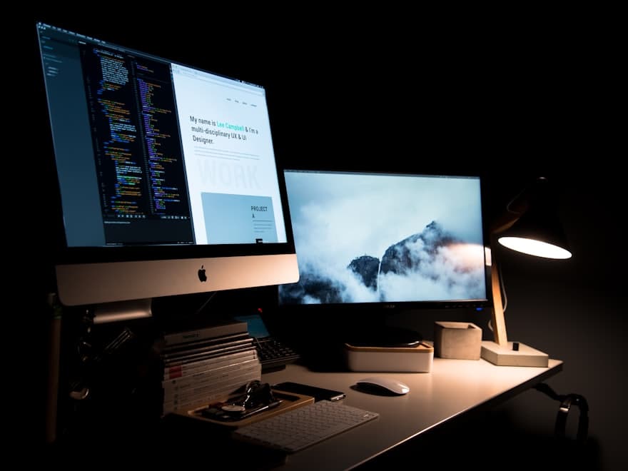 A dark desk with two monitors showing code, lit by a single lamp