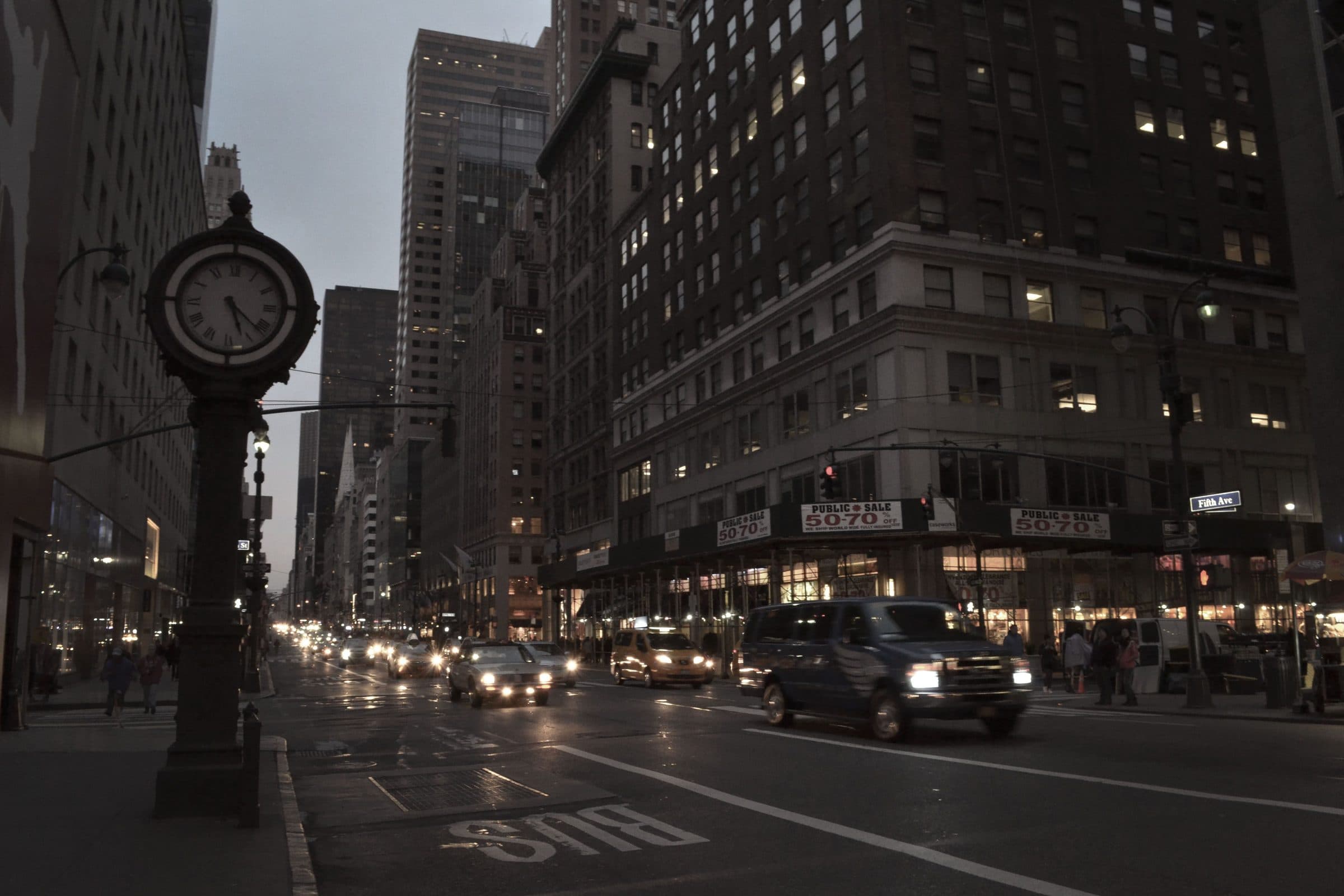 Manhattan street corner at dusk with a street clock and passing traffic